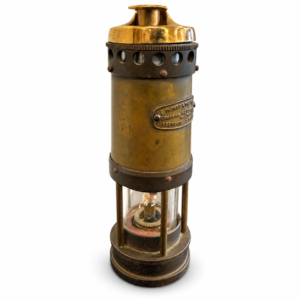Antique-style brass miner’s safety lamp with cylindrical body, glass chamber, dark metal accents, and aged patina.